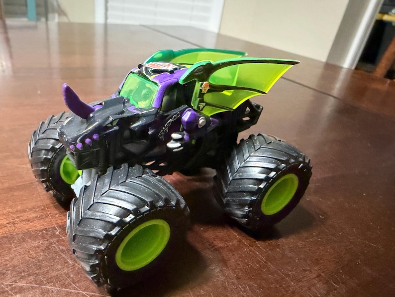 May include: A black, purple, and green monster truck toy with large, black tyres and green rims. The truck features a dragon-themed design with a purple horn and green wing-like accents. The word "Dragonoidz" is visible on the side.