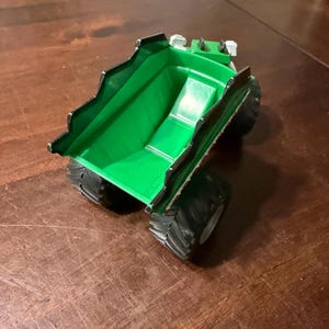 May include: A green toy dump truck with black wheels and a black trim. The truck bed is a bright green color, and the wheels are large and textured. The toy is sitting on a wooden surface.