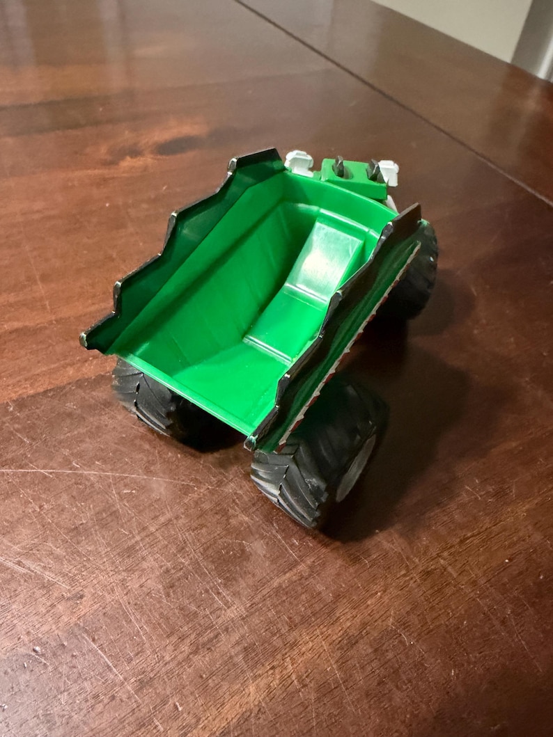 May include: A green toy dump truck with black wheels and a black trim. The truck bed is a bright green color, and the wheels are large and textured. The toy is sitting on a wooden surface.