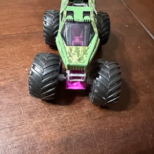May include: A green and camouflage monster truck toy with large black tires. The truck has a purple tinted windshield and a pink chassis. The body of the truck has a camouflage pattern.