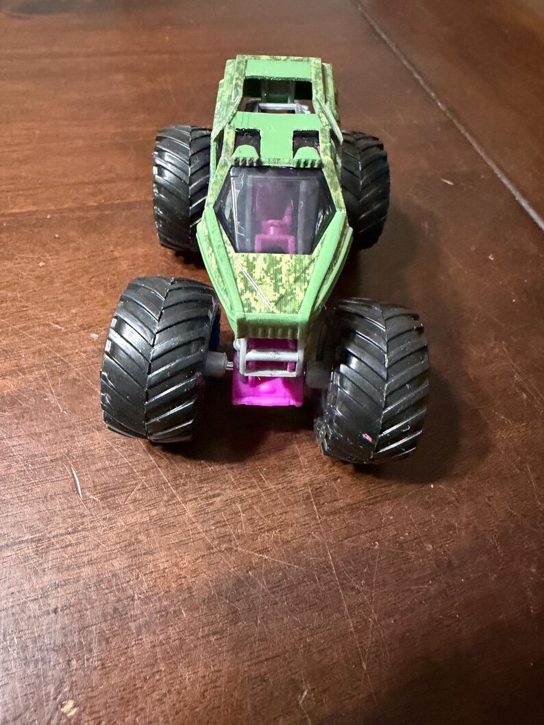 May include: A green and camouflage monster truck toy with large black tires. The truck has a purple tinted windshield and a pink chassis. The body of the truck has a camouflage pattern.
