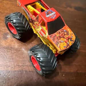 K&ouml;nnte beinhalten: Ein roter und gelber Spielzeug-Monstertruck mit gro&szlig;en schwarzen Reifen und roten Felgen. Der Truck tr&auml;gt die Aufschriften "DRAGONOID" und "BAKUGAN" sowie farbenfrohe Grafiken.