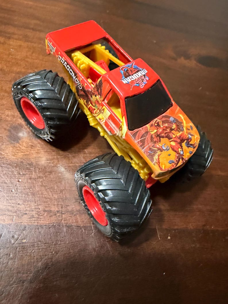 K&ouml;nnte beinhalten: Ein roter und gelber Spielzeug-Monstertruck mit gro&szlig;en schwarzen Reifen und roten Felgen. Der Truck tr&auml;gt die Aufschriften "DRAGONOID" und "BAKUGAN" sowie farbenfrohe Grafiken.