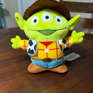 May include: A plush green alien toy with three eyes, wearing a brown cowboy hat, a yellow shirt with red plaid, a vest with black and white cow print, blue pants, and brown boots. The toy is a character from the movie Toy Story.