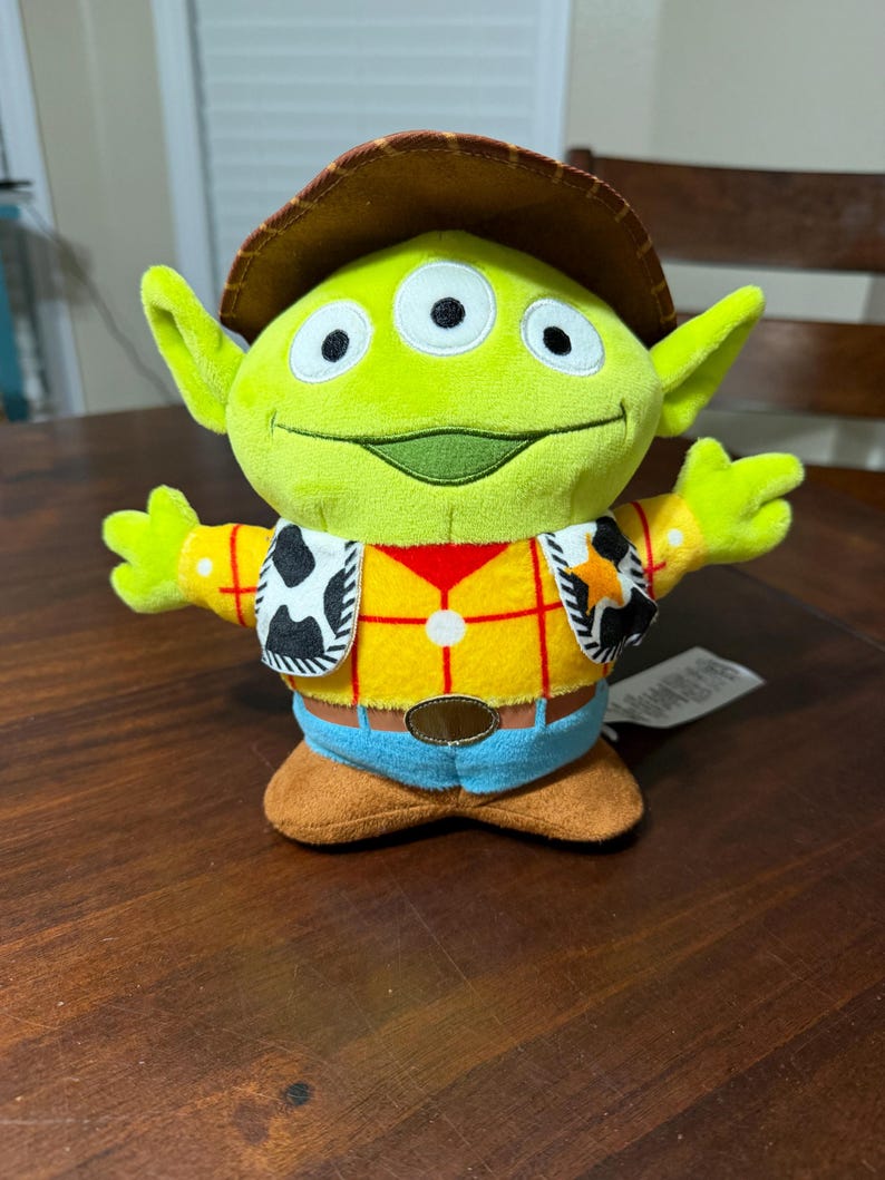 May include: A plush green alien toy with three eyes, wearing a brown cowboy hat, a yellow shirt with red plaid, a vest with black and white cow print, blue pants, and brown boots. The toy is a character from the movie Toy Story.