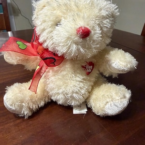 May include: A cream-colored teddy bear with a red nose and a red ribbon bow. The bear has a heart-shaped patch that says "Hug Me". The plush toy is sitting on a wooden surface.