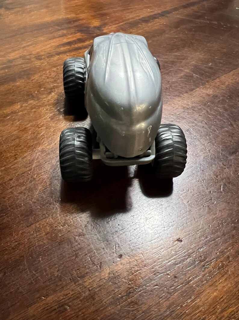May include: A silver toy monster truck with large black wheels. The truck has a unique, streamlined design.