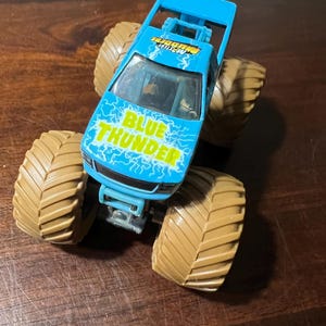 May include: A blue monster truck toy with the words "BLUE THUNDER" in white and yellow lightning bolt graphics on the hood. The truck has large tan tyres and a blue roll cage.