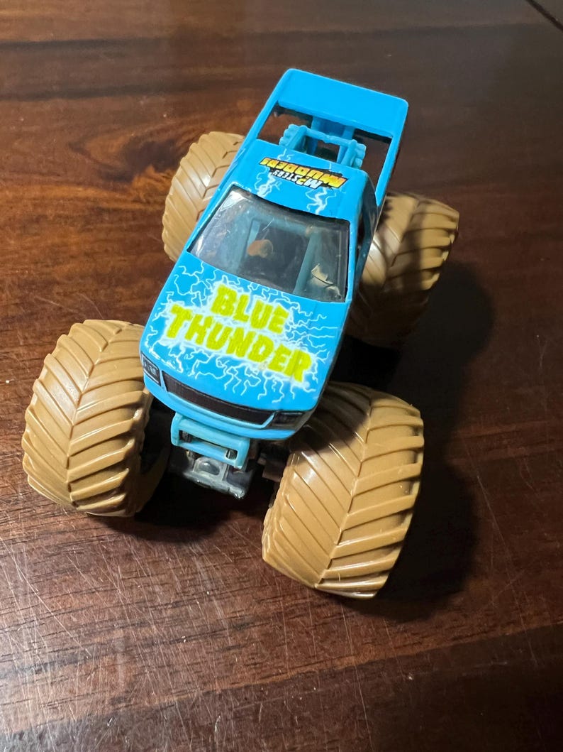 May include: A blue monster truck toy with the words "BLUE THUNDER" in white and yellow lightning bolt graphics on the hood. The truck has large tan tyres and a blue roll cage.