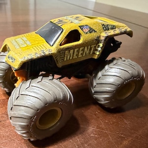 May include: A yellow toy monster truck with large gray tires. The truck has the word "MEENTS" printed on the side and "WORLD CHAMPION" on the top. The truck has a black windshield and a small driver figure.
