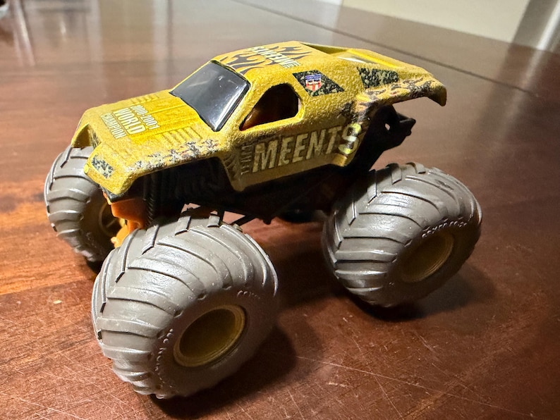 Peut inclure: Un camion monstre jouet jaune avec de gros pneus gris. Le camion porte l'inscription "MEENTS" sur le c&ocirc;t&eacute; et "WORLD CHAMPION" sur le dessus. Le camion a un pare-brise noir et une petite figurine de pilote.