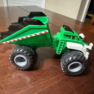 May include: A green and black toy dump truck with large black tires. The truck bed is green with a black top and red and white stripes. The cab is green and white.