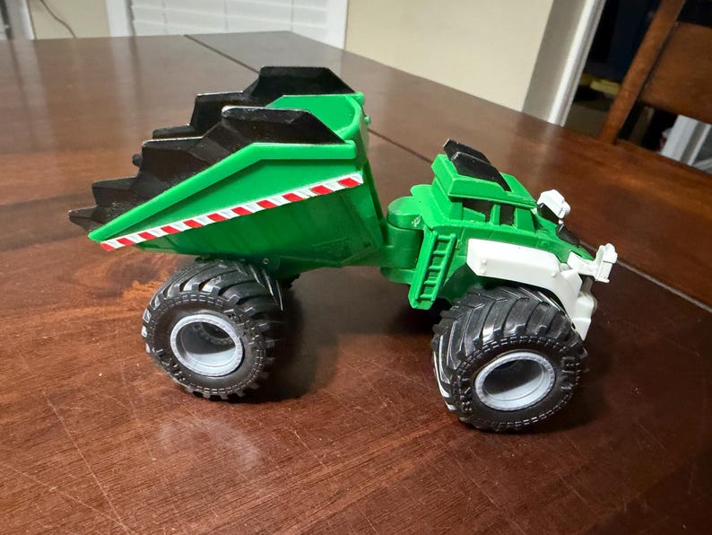 May include: A green and black toy dump truck with large black tires. The truck bed is green with a black top and red and white stripes. The cab is green and white.