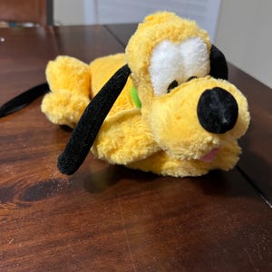以下が含まれることがあります： 黒い耳と鼻を持つ黄色の犬、プルートのぬいぐるみ。おもちゃは白い顔、緑の襟、ピンクの舌をしています。おもちゃは木製の表面に横たわっています。