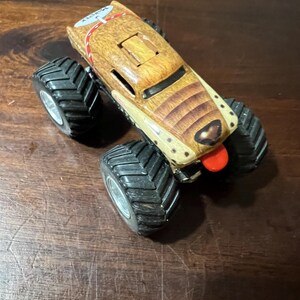 May include: A toy monster truck with a brown and tan body, designed to resemble a dog, with a red tongue. It has large black tyres with silver rims. The top of the truck has the text "MAX CITY".