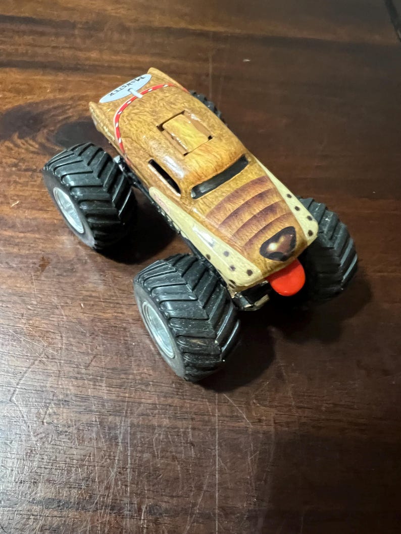 May include: A toy monster truck with a brown and tan body, designed to resemble a dog, with a red tongue. It has large black tyres with silver rims. The top of the truck has the text "MAX CITY".