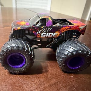 Pode incluir: Um brinquedo de caminh&atilde;o monstro Wild Side com pneus pretos grandes e aros roxos. A carroceria do caminh&atilde;o &eacute; preta com detalhes em laranja e roxo, e as palavras "WILD SIDE" s&atilde;o impressas na lateral.