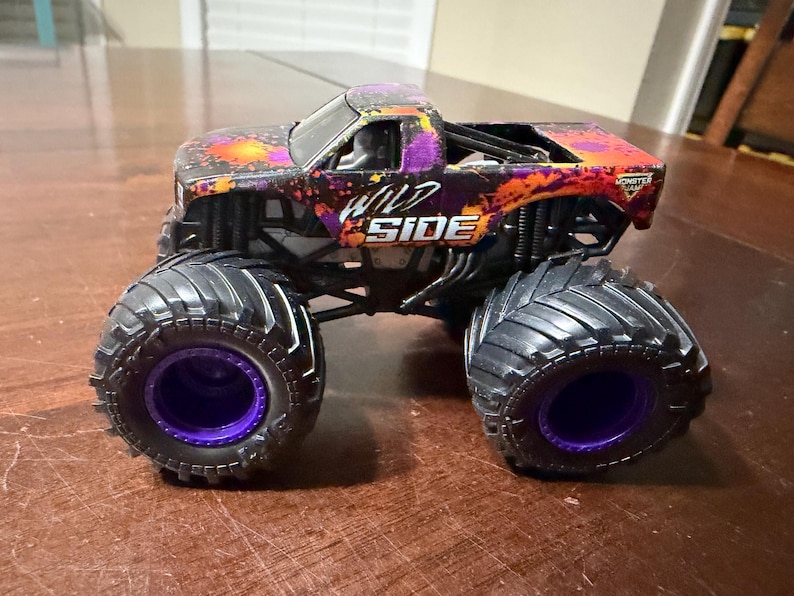 Pode incluir: Um brinquedo de caminh&atilde;o monstro Wild Side com pneus pretos grandes e aros roxos. A carroceria do caminh&atilde;o &eacute; preta com detalhes em laranja e roxo, e as palavras "WILD SIDE" s&atilde;o impressas na lateral.