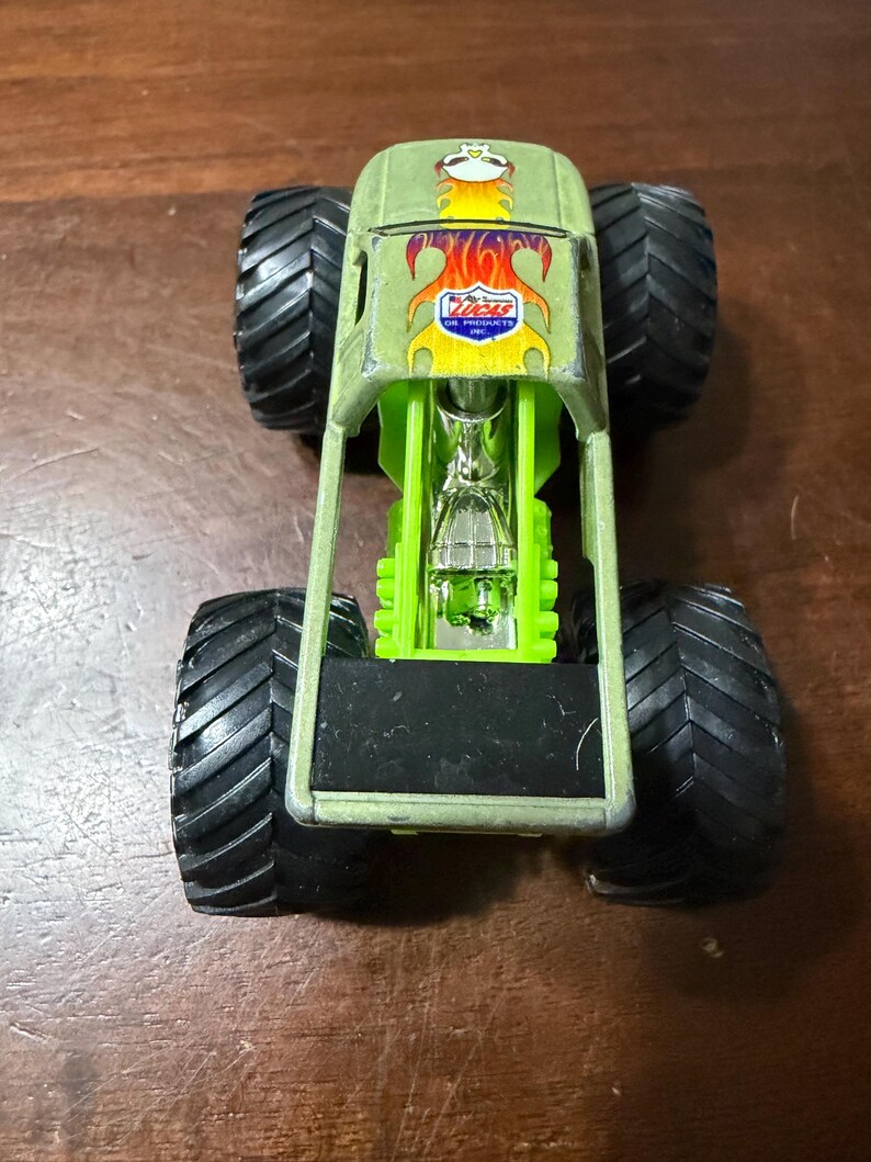 Puede incluir: Un juguete de cami&oacute;n monstruo verde con neum&aacute;ticos negros grandes. El cami&oacute;n tiene un marco verde y un motor detallado. La parte delantera del cami&oacute;n presenta un logotipo con el texto "Lucas Oil Products Inc."