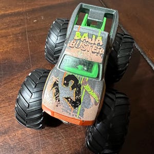 May include: A silver and orange Hot Wheels Baja Buster monster truck toy. The truck has large black tires and the text "BAJA BUSTER" on the roof. The number "3" is on the front of the truck.