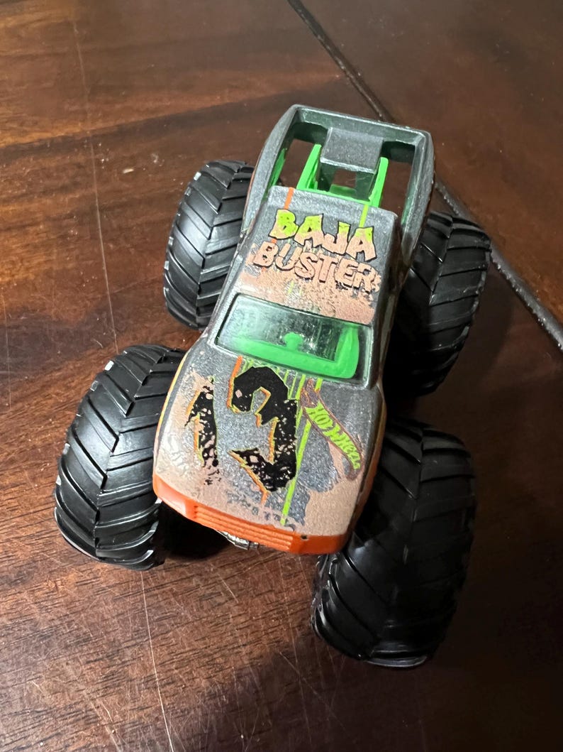 May include: A silver and orange Hot Wheels Baja Buster monster truck toy. The truck has large black tires and the text "BAJA BUSTER" on the roof. The number "3" is on the front of the truck.