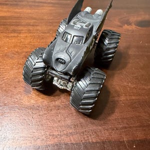 Puede incluir: Un juguete de cami&oacute;n monstruo negro con el tema de Batman con neum&aacute;ticos grandes y negros con banda de rodadura. El cami&oacute;n tiene un s&iacute;mbolo de murci&eacute;lago en la parte delantera y el texto "2 TIME" en la parte superior. El juguete est&aacute; sobre una superficie de madera.