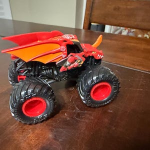May include: A red and orange monster truck toy with a dragon design. The toy has large black tires with red rims and a detailed chassis. The truck features a dragon head and wing design.