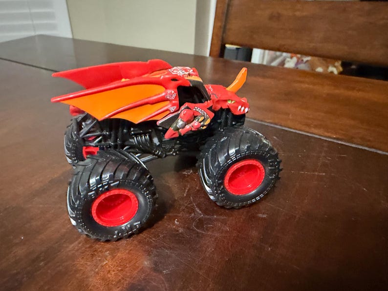 May include: A red and orange monster truck toy with a dragon design. The toy has large black tires with red rims and a detailed chassis. The truck features a dragon head and wing design.