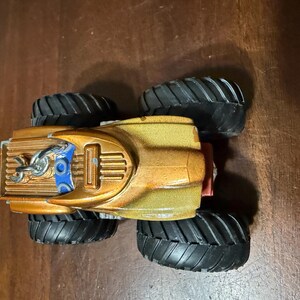 May include: A toy monster truck with a gold and bronze body, black tyres, and blue accents. The truck has a detailed design with a chain and a red rear bumper. The tyres are large and have a deep tread pattern.