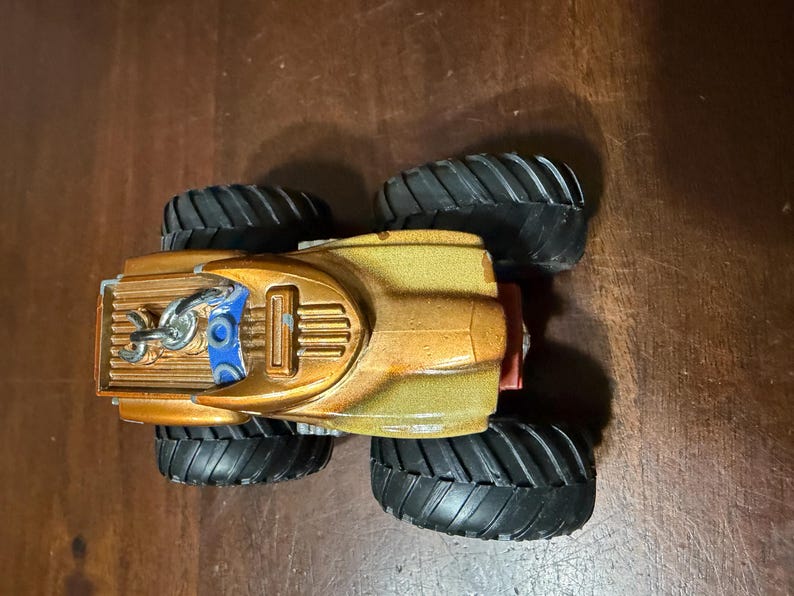 May include: A toy monster truck with a gold and bronze body, black tyres, and blue accents. The truck has a detailed design with a chain and a red rear bumper. The tyres are large and have a deep tread pattern.