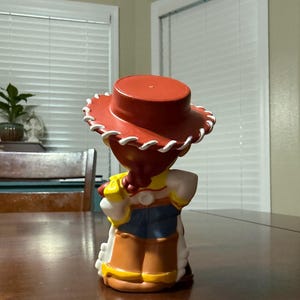 May include: A figurine of a character from the animated film Toy Story, viewed from the back. The figure wears a red hat with white trim, a yellow shirt, a white blouse, a blue vest, and brown chaps. The figure is standing on a wooden table.