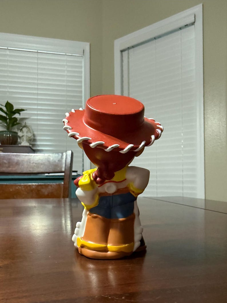 May include: A figurine of a character from the animated film Toy Story, viewed from the back. The figure wears a red hat with white trim, a yellow shirt, a white blouse, a blue vest, and brown chaps. The figure is standing on a wooden table.