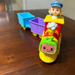 May include: A colorful toy train with a smiling face, a conductor figure, and two attached cars. The train is primarily red, yellow, and green, with blue and purple cars. The conductor wears a blue cap.