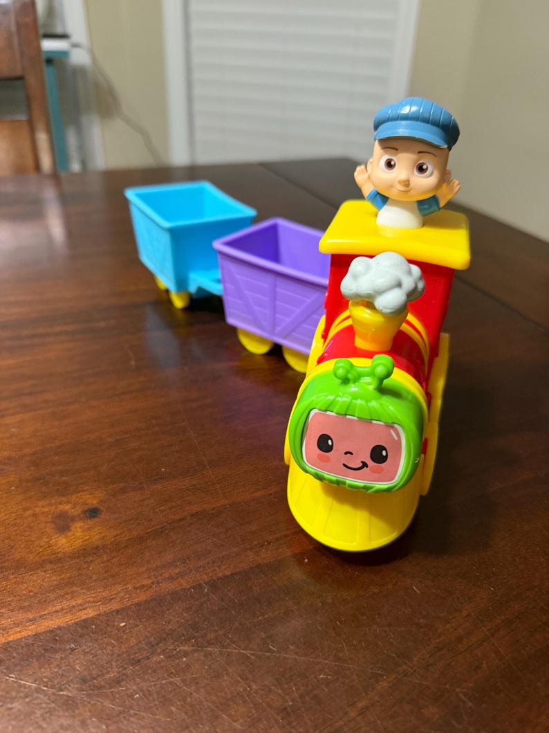 May include: A colorful toy train with a smiling face, a conductor figure, and two attached cars. The train is primarily red, yellow, and green, with blue and purple cars. The conductor wears a blue cap.
