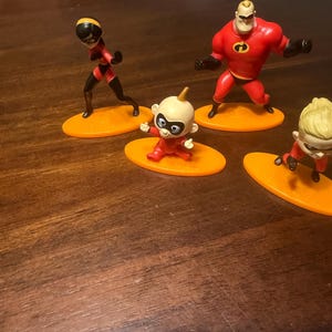 May include: Four figurines of characters from The Incredibles movie. The figures are on orange oval bases. The characters include Mr. Incredible in a red suit, Elastigirl in a red and black suit, and two versions of Jack-Jack in red suits.