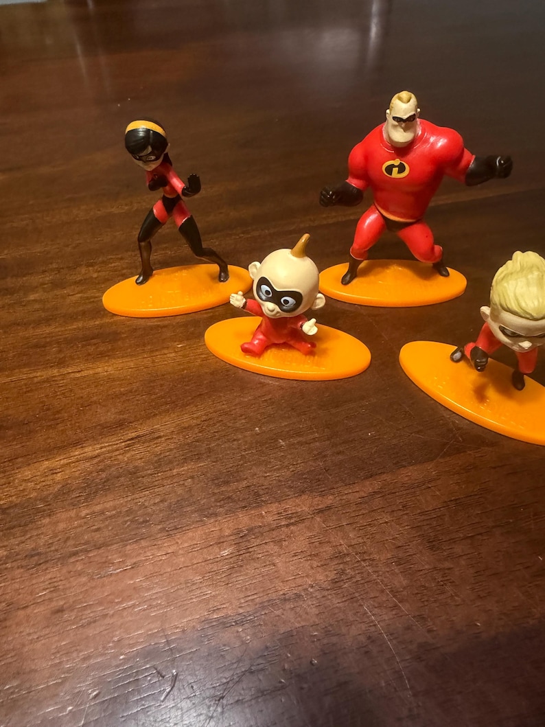 May include: Four figurines of characters from The Incredibles movie. The figures are on orange oval bases. The characters include Mr. Incredible in a red suit, Elastigirl in a red and black suit, and two versions of Jack-Jack in red suits.