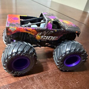 Pode incluir: Um monster truck de brinquedo com pneus pretos grandes e aros roxos. A carroceria do caminh&atilde;o &eacute; preta com detalhes roxos e laranja, e as palavras "WILD SIDE" s&atilde;o vis&iacute;veis na lateral. &Eacute; um ve&iacute;culo Monster Jam.