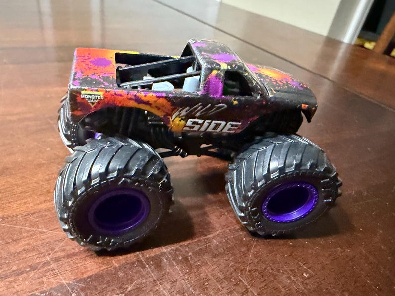 Pode incluir: Um monster truck de brinquedo com pneus pretos grandes e aros roxos. A carroceria do caminh&atilde;o &eacute; preta com detalhes roxos e laranja, e as palavras "WILD SIDE" s&atilde;o vis&iacute;veis na lateral. &Eacute; um ve&iacute;culo Monster Jam.