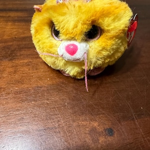 May include: A plush, round cat toy with yellow fur, large black eyes, and a pink nose. It has a pink glitter bow on its head and pink whiskers. A red tag is attached to the side.