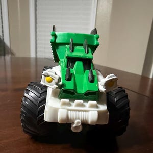 May include: A toy truck with a green cab and white chassis. The truck has large black tires and yellow accents on the front. The toy is designed with a rugged, industrial aesthetic.