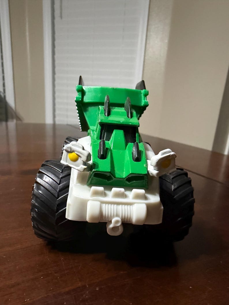 May include: A toy truck with a green cab and white chassis. The truck has large black tires and yellow accents on the front. The toy is designed with a rugged, industrial aesthetic.