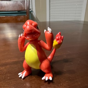 May include: A red and yellow figurine of a Charmander character, with white accents on its claws. The figure is in a dynamic pose, with its arms raised and tail up. The figure has a yellow belly and a flame on its tail.