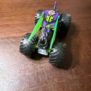 May include: A toy monster truck with large black tyres. The truck has a purple body with green accents and a logo that says "AKURA 20". The truck is sitting on a wooden surface.