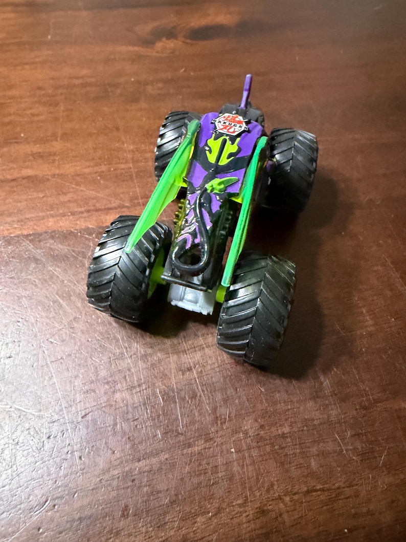 May include: A toy monster truck with large black tyres. The truck has a purple body with green accents and a logo that says "AKURA 20". The truck is sitting on a wooden surface.