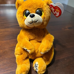 May include: A plush orange teddy bear with large, glittery eyes and a black nose and mouth. The bear has a tag attached to its ear. The bear is sitting on a dark brown surface.