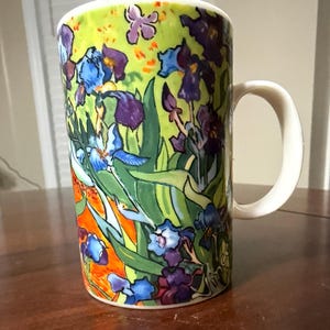 May include: A ceramic coffee mug featuring a colorful floral design with irises in shades of blue and purple, set against a green and orange background. The mug has a white handle and a white rim.