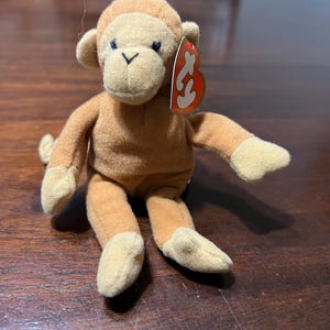 May include: A plush monkey toy with light brown fur, tan hands and feet, and a tan face. The monkey has black eyes and a red and white tag attached to its ear. The toy is sitting on a wooden surface.