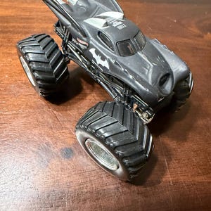 Puede incluir: Un cami&oacute;n monstruo Hot Wheels negro con el tema de Batman. El cami&oacute;n de juguete presenta neum&aacute;ticos negros grandes, un s&iacute;mbolo de murci&eacute;lago en el lateral y un dise&ntilde;o en forma de ala en la parte superior. El veh&iacute;culo est&aacute; sobre una superficie de madera.