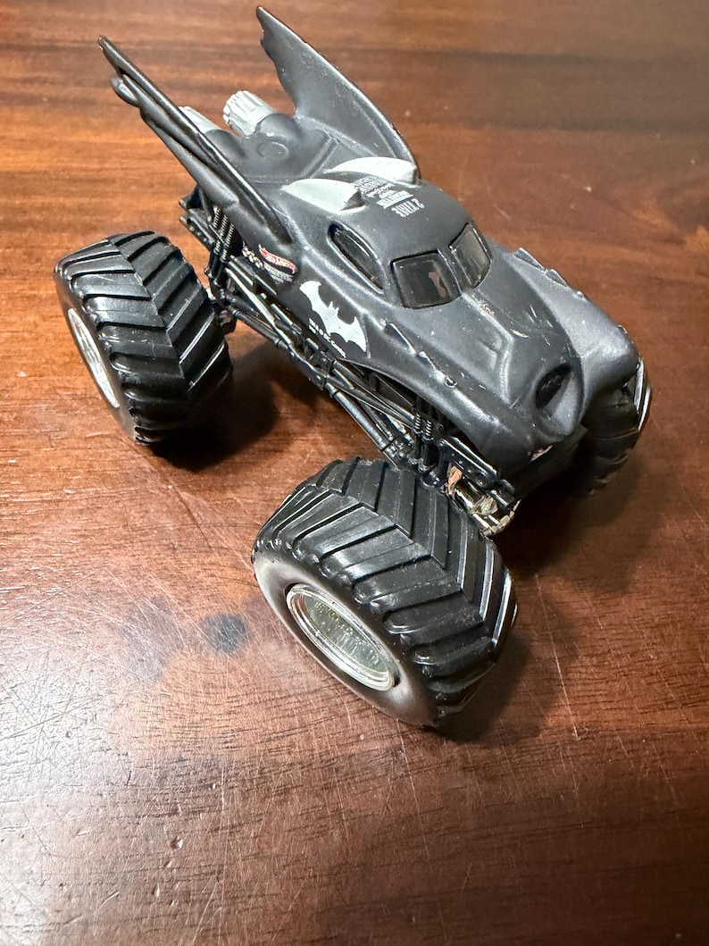 Puede incluir: Un cami&oacute;n monstruo Hot Wheels negro con el tema de Batman. El cami&oacute;n de juguete presenta neum&aacute;ticos negros grandes, un s&iacute;mbolo de murci&eacute;lago en el lateral y un dise&ntilde;o en forma de ala en la parte superior. El veh&iacute;culo est&aacute; sobre una superficie de madera.