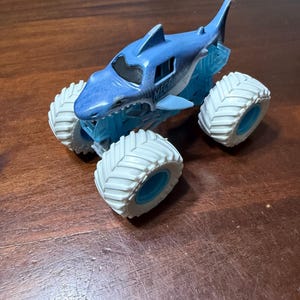 Puede incluir: Un cami&oacute;n monstruo de juguete azul y blanco con un dise&ntilde;o de tibur&oacute;n. El cami&oacute;n tiene neum&aacute;ticos blancos grandes con centros azules y la palabra "MEGAlodon" impresa en el lateral.